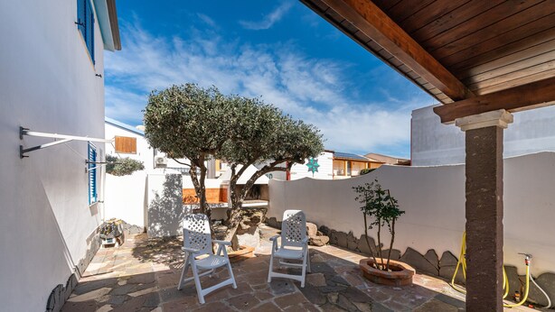 Un patio con suelo de piedra, dos sillas blancas, un árbol y una manguera.