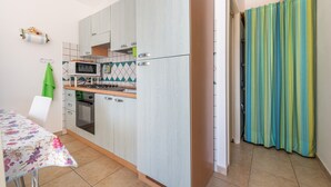 A compact kitchen with a microwave, stove, and oven, a dining table with a floral tablecloth, and a green chair. - Holiday Home "Sottotorre 2" close to the Sea with Private Garden & A/C (Calasetta)