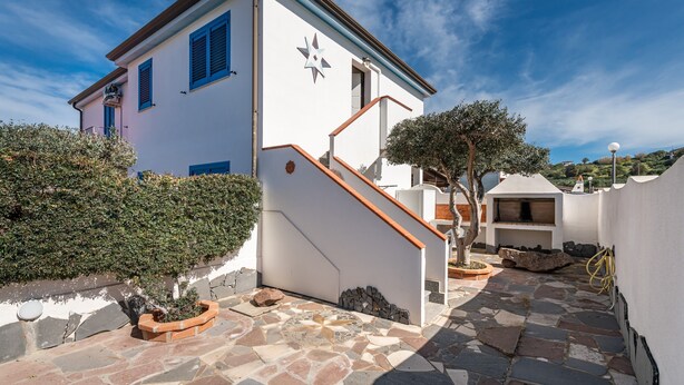 A white building with blue shutters, a star-shaped window, and a stone staircase leading to a covered area with a barbecue. - Holiday Home "Sottotorre 2" close to the Sea with Private Garden & A/C (Calasetta)