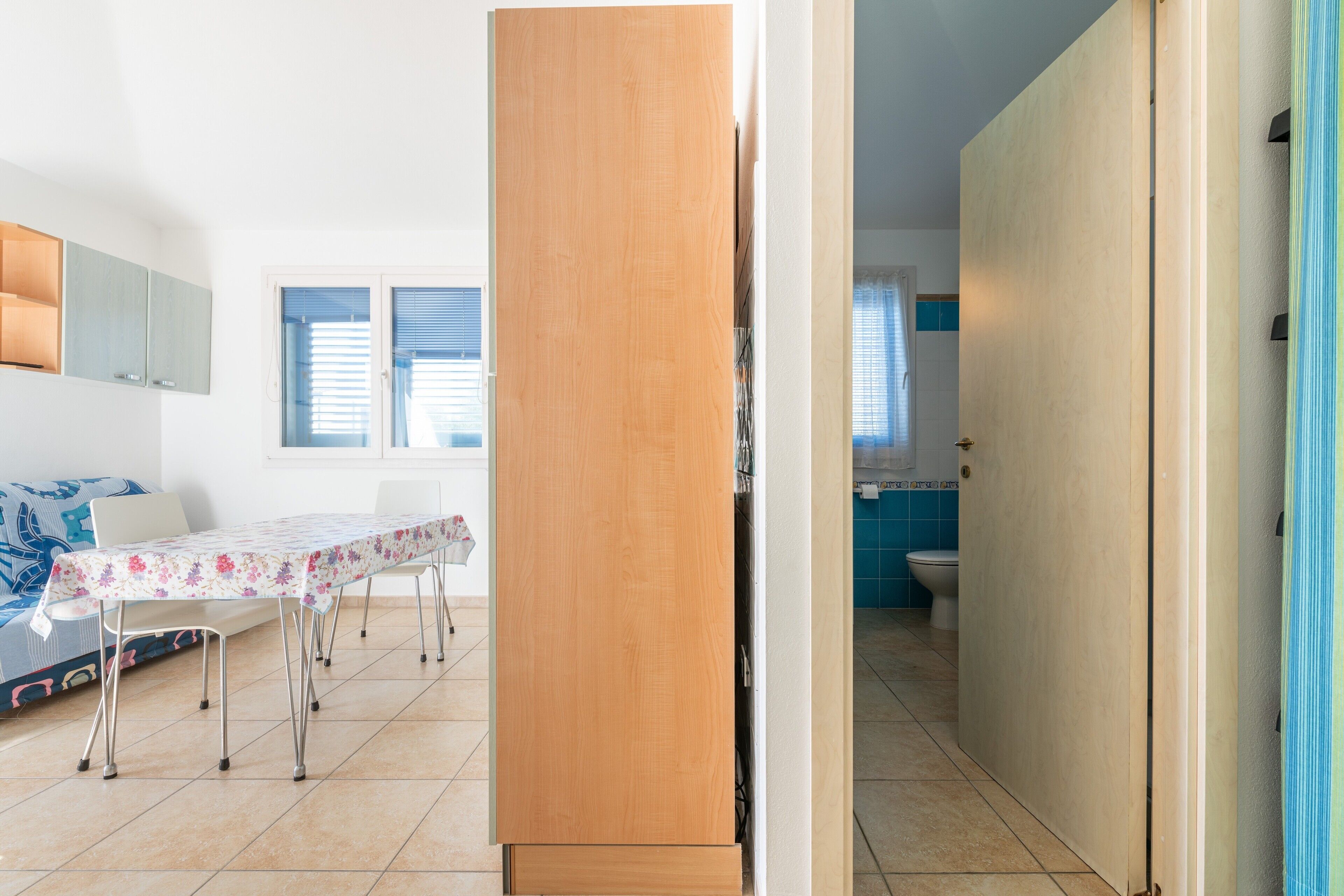 Ein Zimmer mit Esstisch, Stühlen, einem Holzkasten, einem Badezimmer mit Toilette und einem Fenster mit Jalousien.