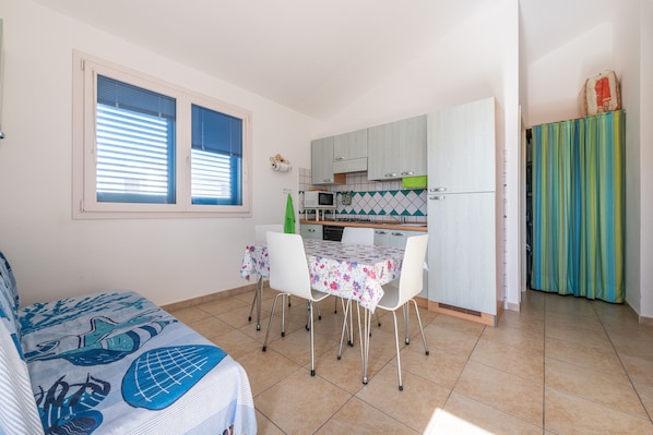 Une cuisine de taille réduite avec des meubles blancs, une table à manger avec des chaises et une fenêtre avec des stores.