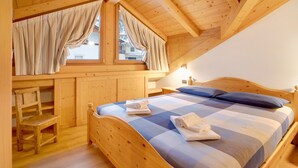 Una camera da letto in legno con un letto, una sedia, una finestra con tende e vista su una casa.