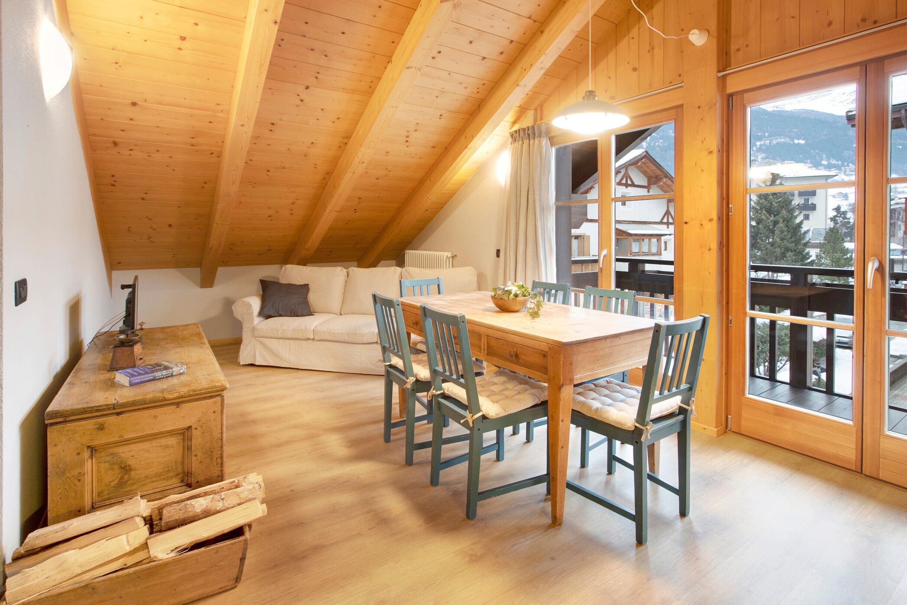 A cozy room with a wooden ceiling, a dining table with chairs, a sofa, and a view of mountains through large windows.
