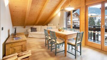 A cozy room with a wooden ceiling, a dining table with chairs, a sofa, and a view of mountains through large windows.