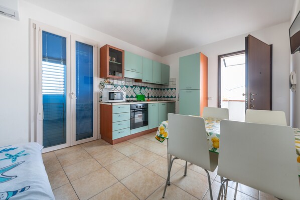 Une cuisine compacte équipée d’un four à micro-ondes, d’une cuisinière et de placards, un coin repas avec une table et des chaises, et une porte-fenêtre coulissante donnant sur un balcon.