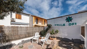 A courtyard with stone flooring, two white chairs, a wooden fence, and a wall with the text 'TILLA DEI'.