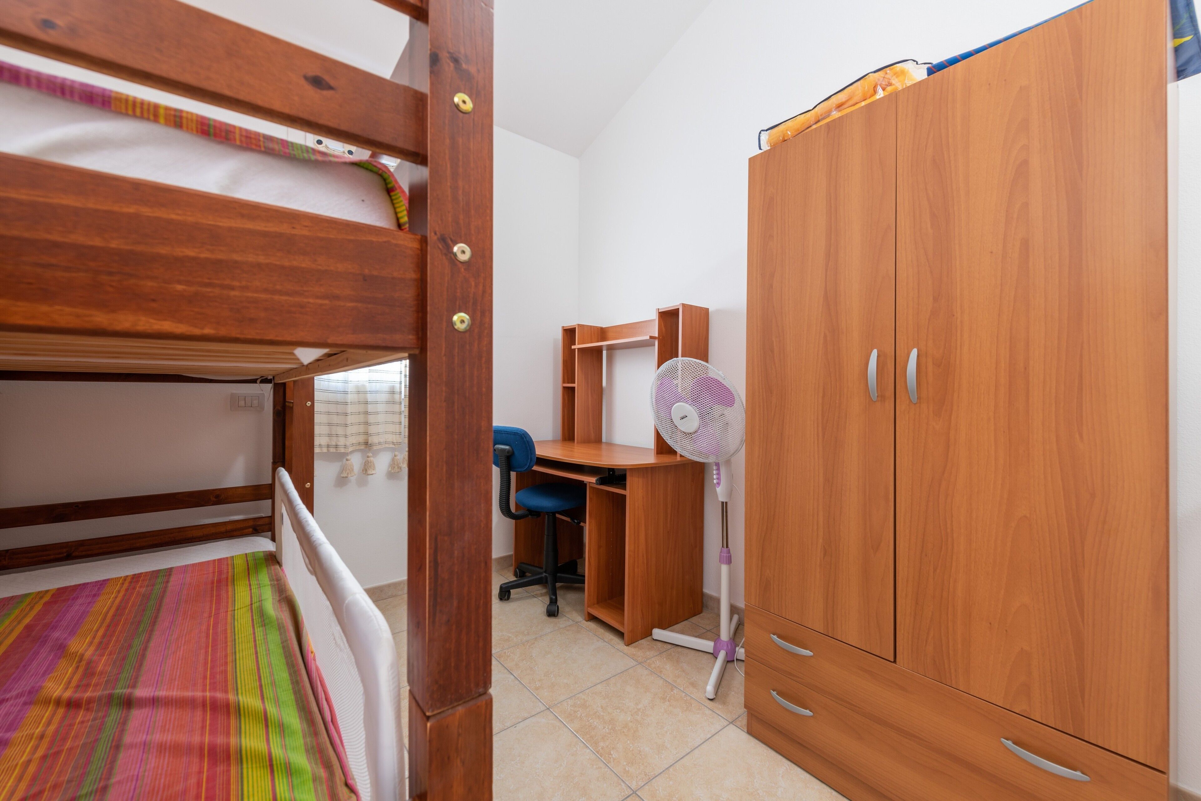 A bedroom with a bunk bed, a desk with a chair, a wardrobe, and a standing fan.