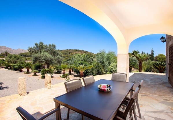 An outdoor patio with a table and chairs, surrounded by stone pillars and a view of a hilly landscape with trees.