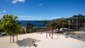 Ein Spielplatz mit Schaukeln und Rutsche, eine Bank und Blick auf das Meer.