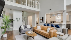A spacious living area with a high ceiling, a balcony, a kitchen with bar seating, and a staircase leading to an upper level.