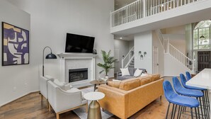 A spacious living room with a fireplace, a sofa, a coffee table, and a TV. There is a staircase leading to an upper level.