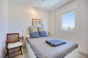 Un dormitorio con cama, una silla, una mesita de noche, un cuadro enmarcado y una ventana con vistas.