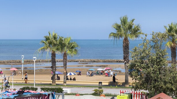 A beach with palm trees, umbrellas, and people. - Apartment “Chipiona, Cruz del Mar 1st Line” with Sea Views, Terrace, Parking & Wi-Fi (Chipiona)