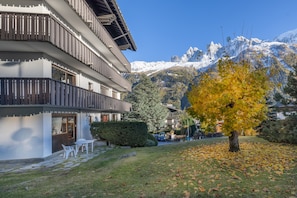 A multi-story building with balconies, a tree with yellow leaves, and snow-capped mountains in the background.