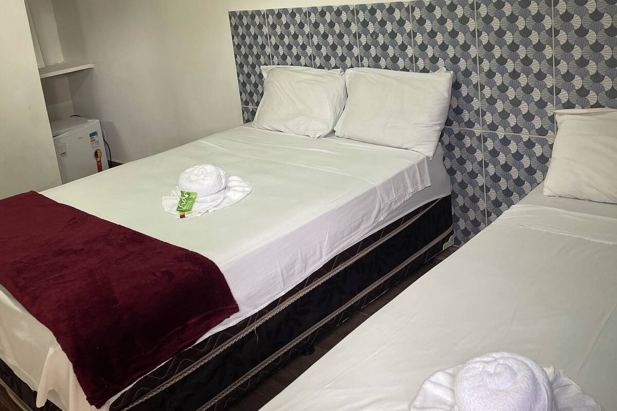 A hotel room with two beds, each with white linens and a red blanket. There is a small white cabinet and a wall-mounted heater.