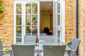 A glass table with wicker chairs, a bottle, and glasses, set against a brick wall with a sliding glass door leading to a room with a piano. - LUXURY DUPLEX IN NEW DEVELOPMENT (London)
