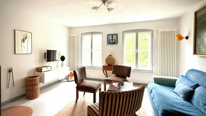 Een woonkamer met een blauwe bank, gestreepte stoelen, een televisie en een raam met gordijnen.