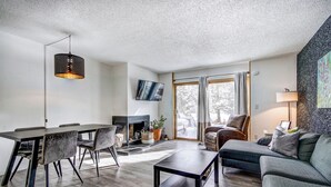 A modern living room with a dining area, a fireplace, and a large window with curtains. - Claimjumper Condo - Hot Tub, Pool, Bus Route, Free Parking (Breckenridge)
