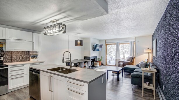 A modern kitchen with white cabinetry, a dishwasher, and a large island with a sink. There is a dining area with a table and chairs, and a living room with a sofa and a coffee table. - Claimjumper Condo - Hot Tub, Pool, Bus Route, Free Parking (Breckenridge)