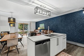 Een moderne keuken met een vaatwasser, een eethoek met een houten tafel en stoelen, en een zithoek met een bank en een schilderij aan de muur.