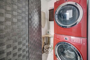 A laundry room with a red front-loading washer and dryer, a wooden shelf with a kettle, and a woven mat on the wall.