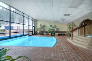 An indoor swimming pool with a tiled floor, large windows, and a stone wall with a staircase leading to a hot tub area.