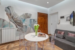Un salon moderne avec une table ronde blanche, des chaises en plastique transparent, un canapé gris et une grande fresque abstraite sur le mur.