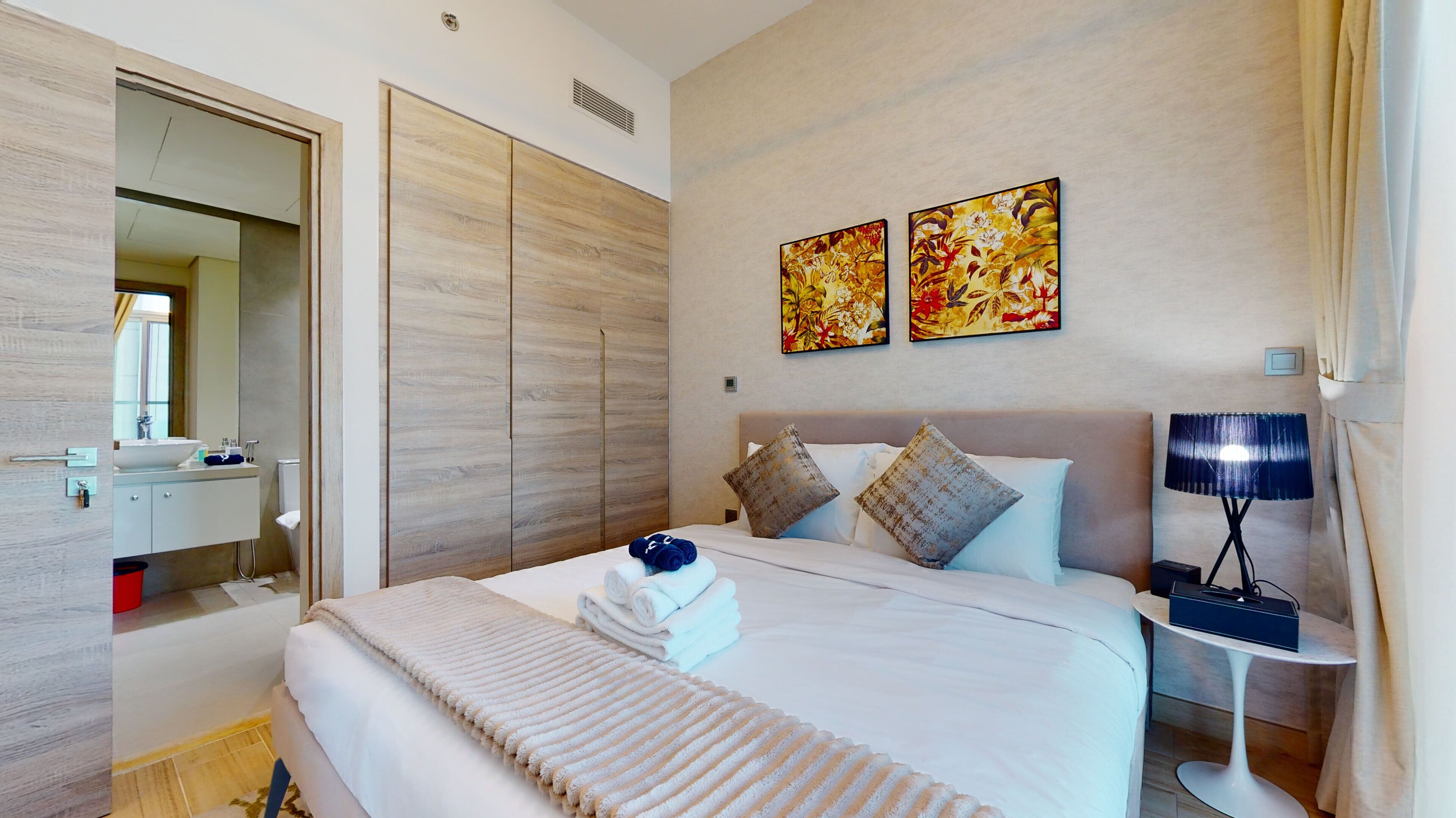 A modern hotel room with a large bed, a bedside table with a lamp, a mirror, a wardrobe, and two floral paintings on the wall.