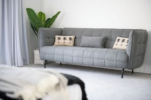 A grey sofa with two matching cushions, a white basket, and a large green plant in a room with white walls. - Whitsunday Holiday Home (Cannonvale)