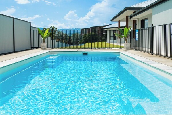 A rectangular swimming pool with clear blue water, surrounded by a black metal fence, and a building with a white facade in the background.