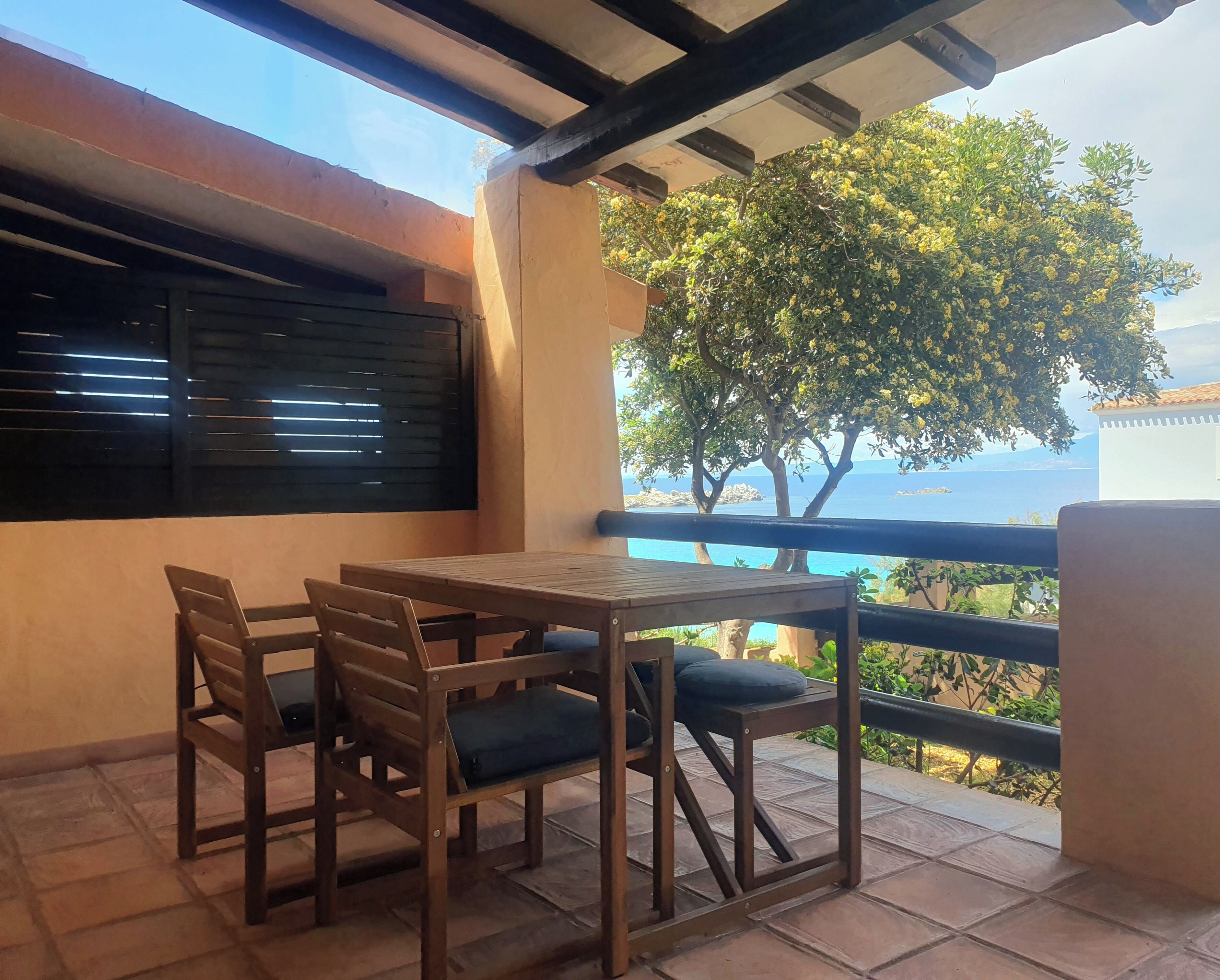 Een balkon met een houten tafel en stoelen, uitzicht op de zee, en een boom met gele bloemen.