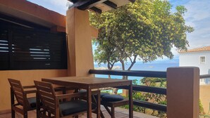 Ett träbord och några stolar står på en terrass med kakelplattor, och härifrån har man utsikt över havet och ett träd.