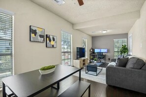 A modern living room with a sofa, a coffee table, and a TV. There are two windows with blinds, and a ceiling fan is visible.