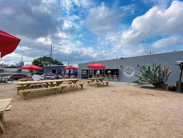 An outdoor patio area with wooden picnic tables and red umbrellas, a grey building with a logo, and string lights.
