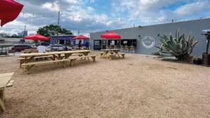 An outdoor seating area with wooden tables and red umbrellas, a grey building with a logo, and a city skyline in the background. - Entire Apt | Gated Parking | KING Bed | Computer (Houston)