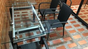 Mesa com tampo de vidro, suportes de metal, cadeiras de palha preta e chão com padrão de tijolos.