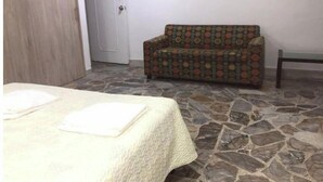 Quarto com piso de pedra, cama com colcha branca, sofá estampado e uma porta.