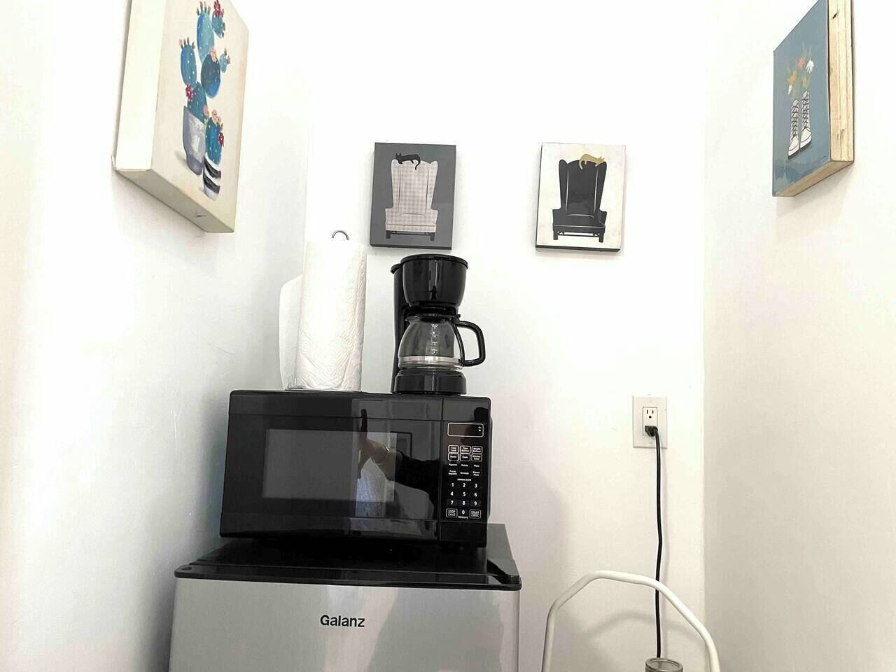 A microwave with a coffee maker on top, a white wall with framed pictures, and a power outlet.