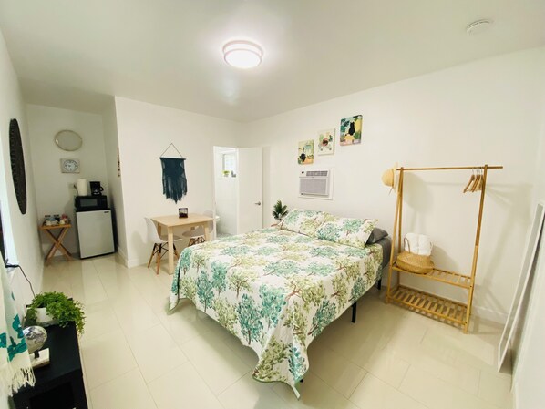 A bedroom with a bed, a desk, a small fridge, a hanging light fixture, and a wall-mounted air conditioner. - Authentic Little Havana • Walk to Calle Ocho (Miami)