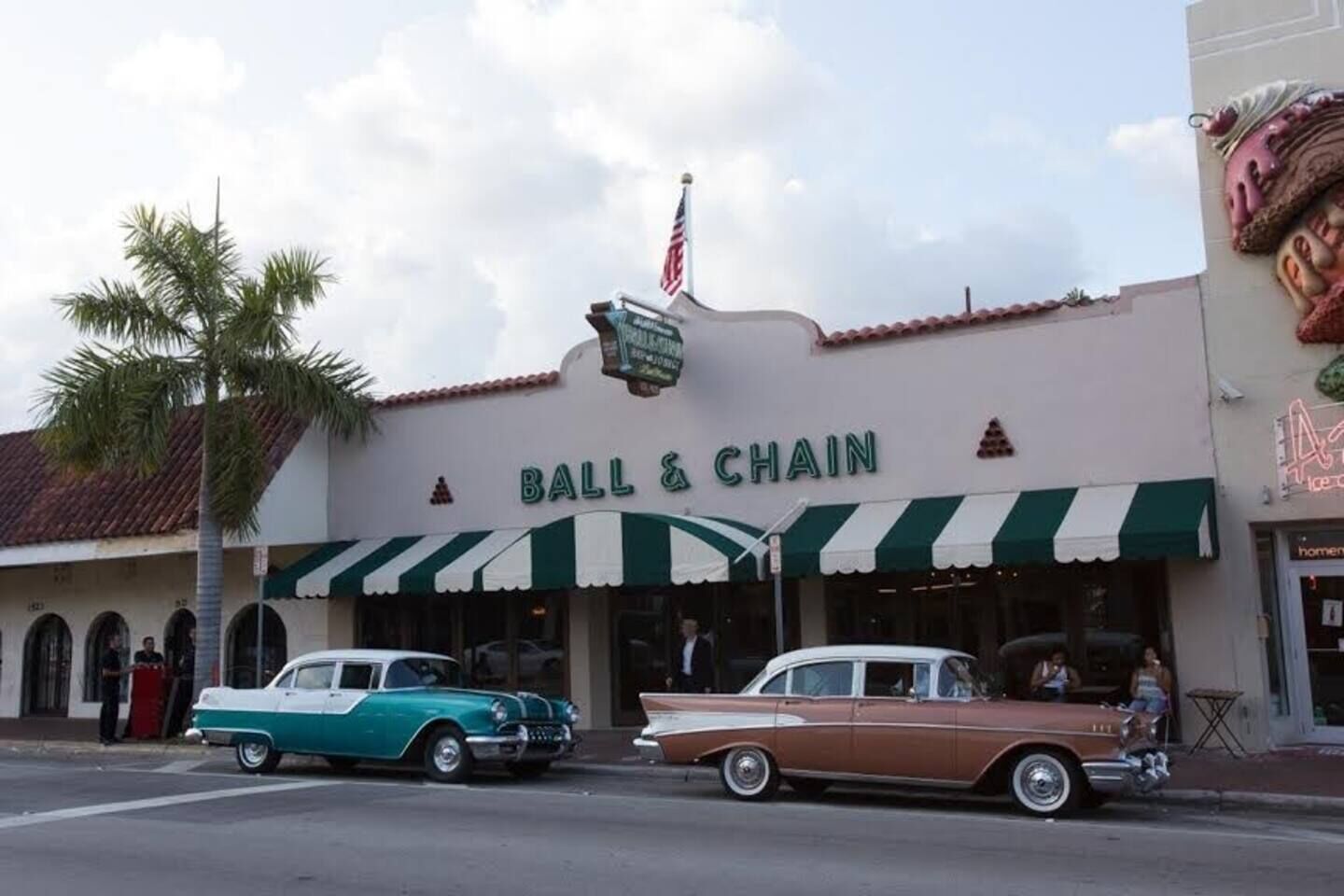 A vintage storefront with a neon sign for 'BALL & CHAIN' and classic cars parked in front.