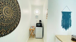 A narrow hallway with a decorative wall piece, a small table with items, a refrigerator, a microwave, and a hanging macrame wall decoration.