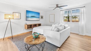A modern bedroom with a bed, a white sofa, a round coffee table, a ceiling fan, a window with curtains, and a painting on the wall. - 3 Queen Beds, 5 min Walk to the Beach - 70 inch Smart TV (Lauderdale-by-the-Sea)