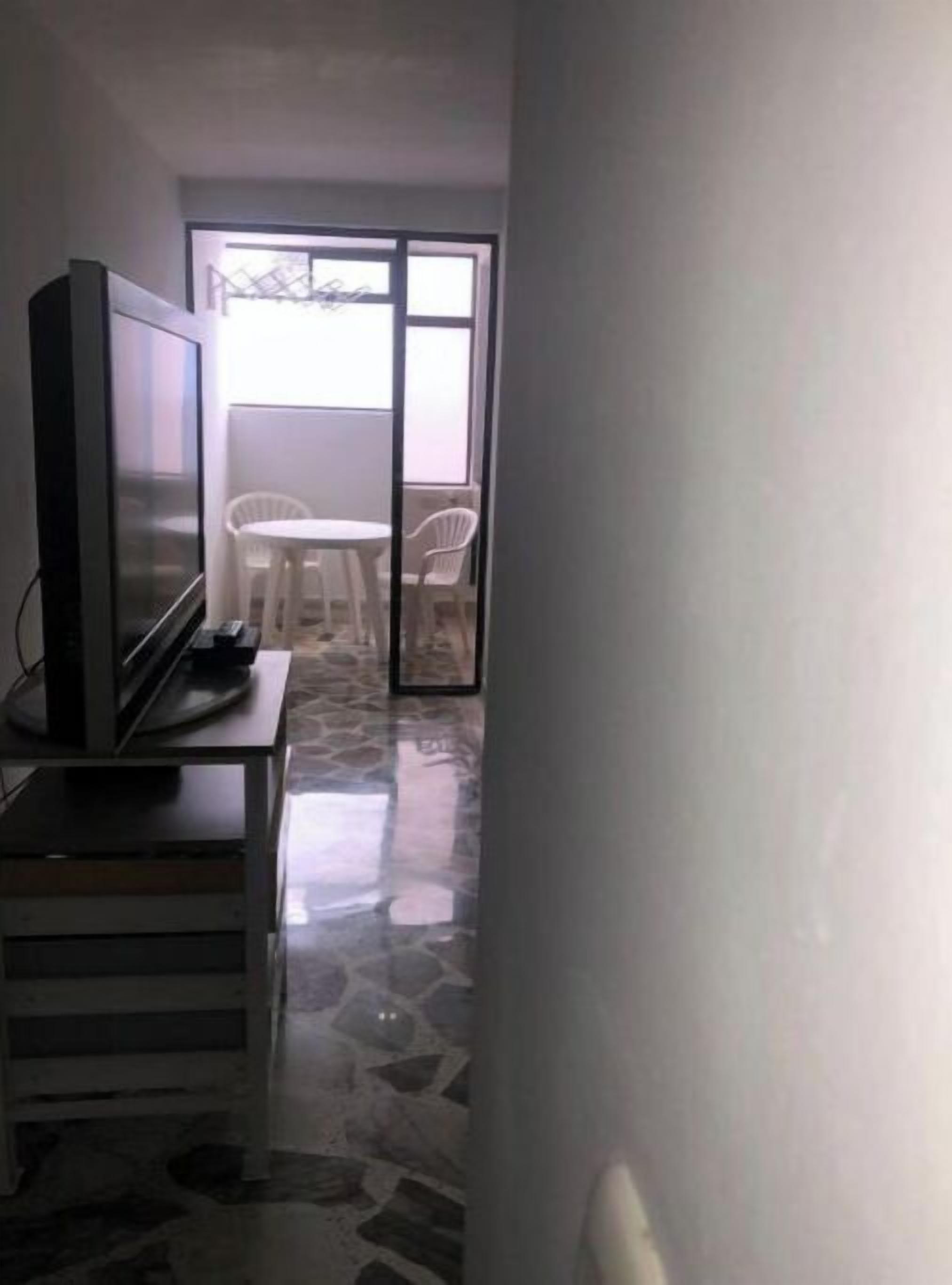 A narrow hallway with a television on a stand, a stone-patterned floor, and a sliding glass door leading to an outdoor area with a table and chairs.