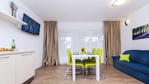A modern living room with a blue sofa, green chairs, a white dining table, and a kitchen area with a white cabinet and a TV on the wall. - Vacation apartment right by the sea-50 m from the beach (Fažana)