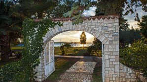 An arched stone gateway with a metal gate, leading to a stone pathway and a grassy area with trees. - Vacation apartment right by the sea-50 m from the beach (Fažana)