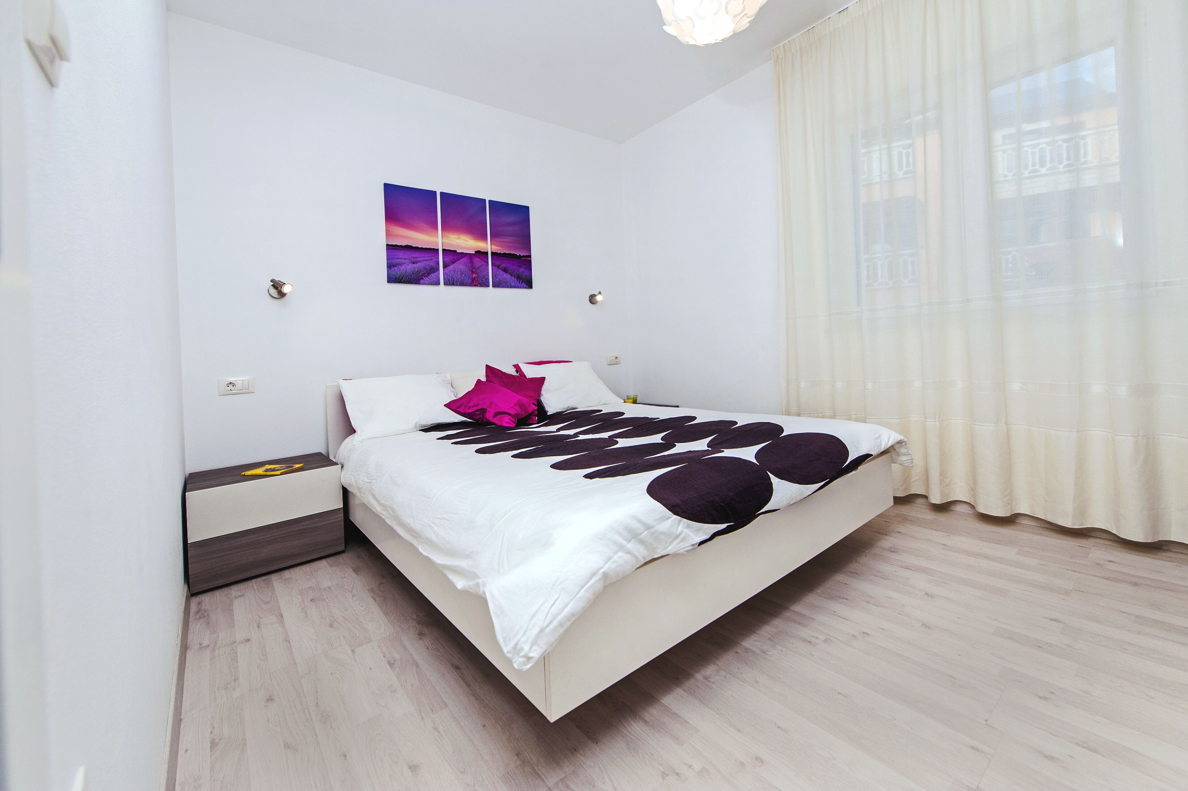 Ein Schlafzimmer mit einem Bett, einem Nachttisch, einer Wandleuchte und einem Fenster mit Vorhängen.