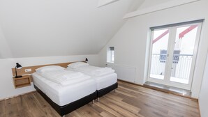 A bedroom with a wooden floor, a bed with white bedding, a nightstand, a window, and a view of the outside.