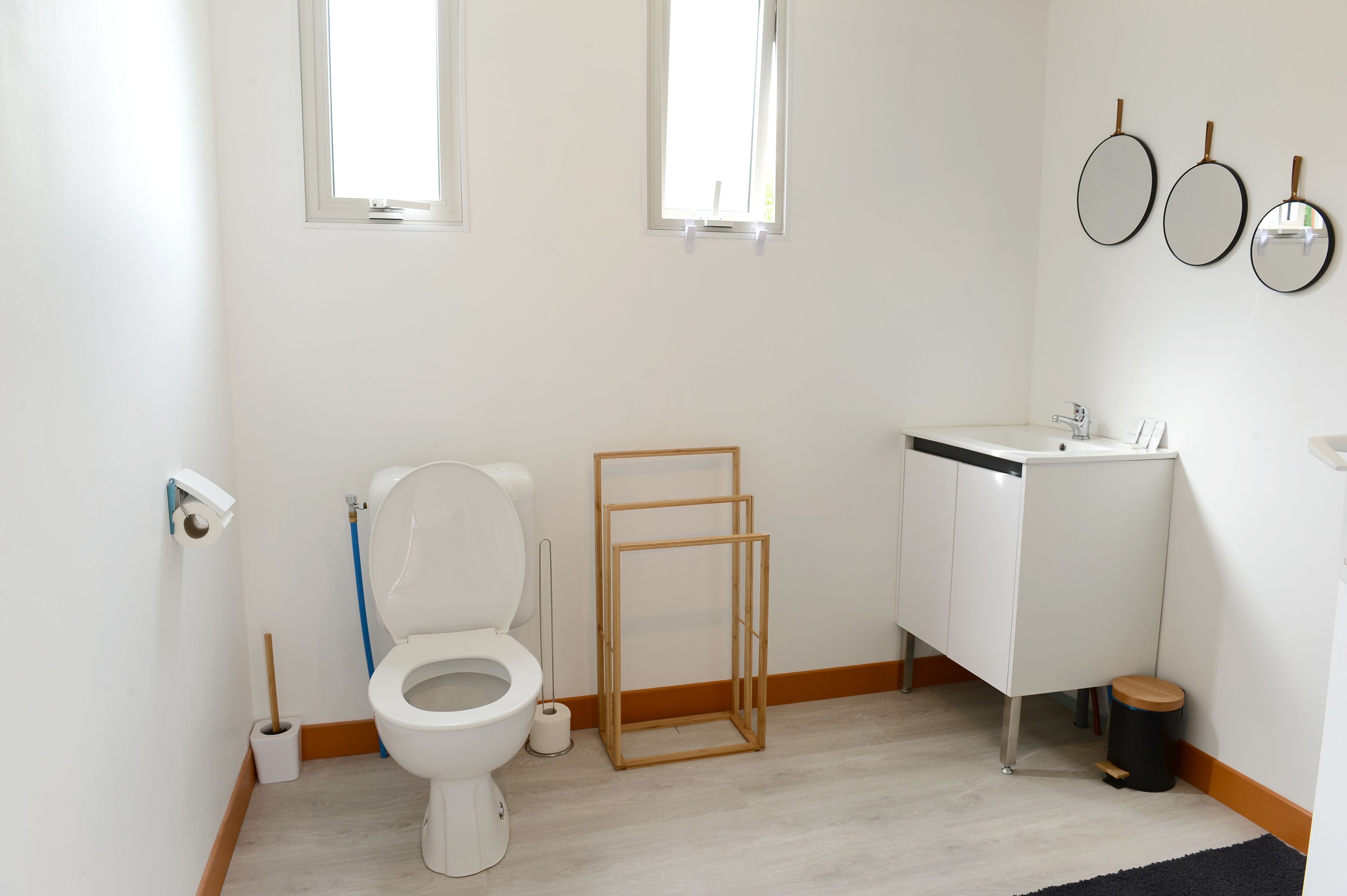 Ein Badezimmer mit Toilette, Waschbecken und Spiegel. Zwei Fenster und ein Holzboden sind vorhanden.