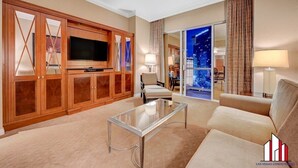 A modern living room with a large flat-screen TV, a glass coffee table, and a sofa. There is a sliding glass door leading to a balcony with a city view.