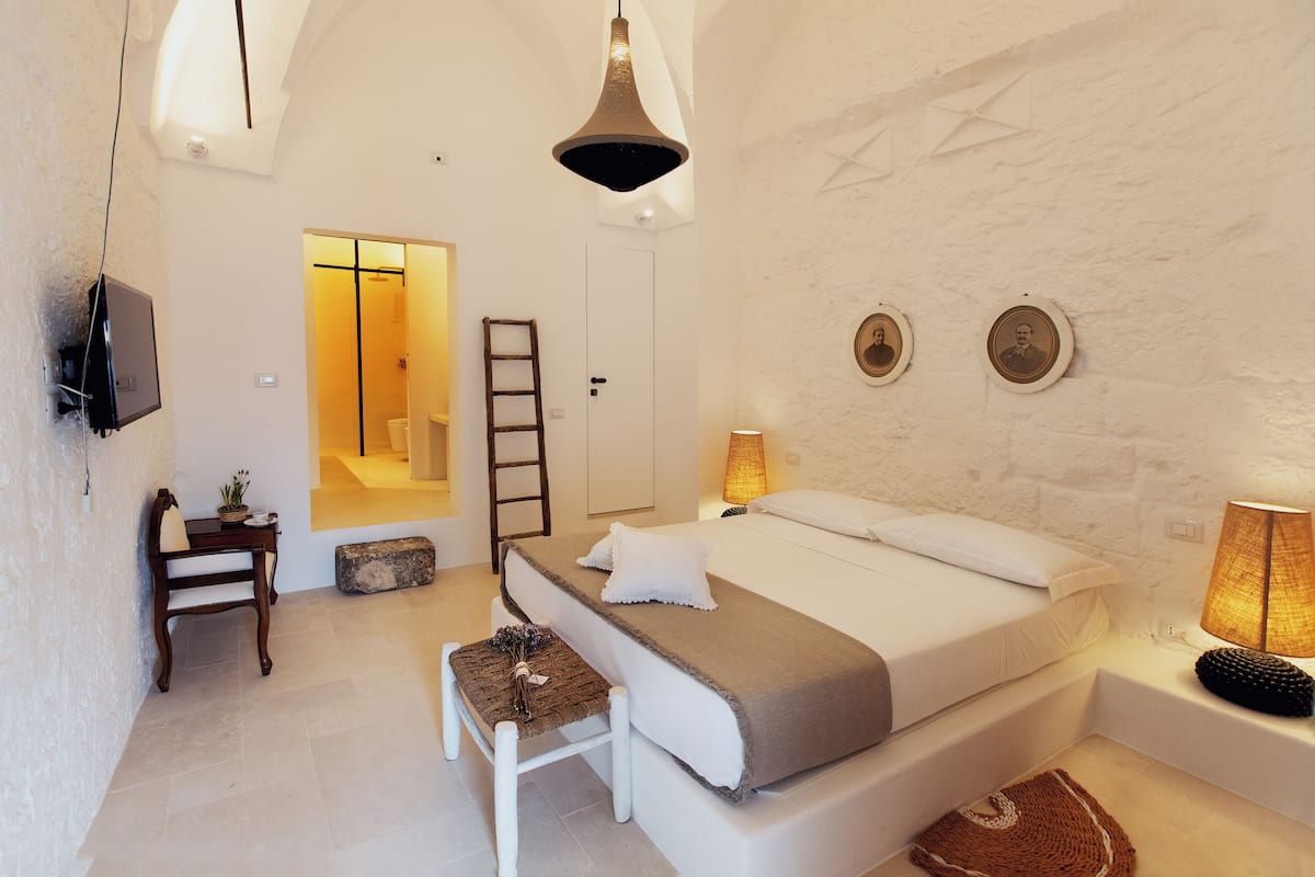 Quarto com cama, abajures, uma mesinha com uma planta, um banco e TV na parede.